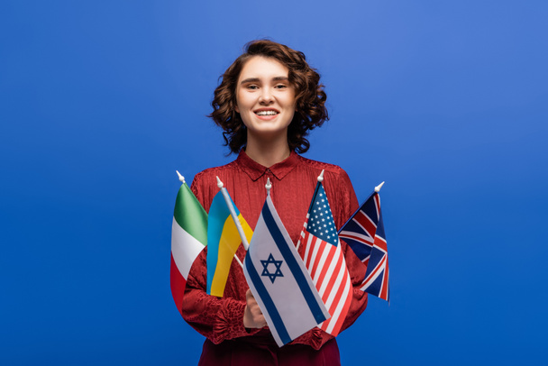 mujer complacida sonriendo a la cámara mientras sostiene banderas de diferentes países aislados en azul - Foto, Imagen