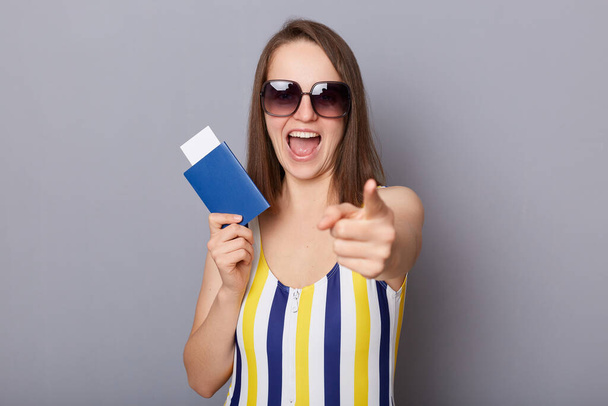 Indoor shot of overjoyed woman wearing swimsuit and sunglasses holding passport and pointing to camera, smiling broadly, inviting you for trip, advertising travel agency, isolated on gray background - Valokuva, kuva