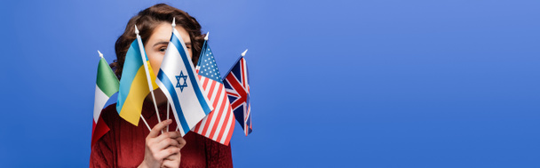 mladá žena zakrývající tvář s různými vlajkami a dívající se na kameru izolovanou na modré, transparentní - Fotografie, Obrázek