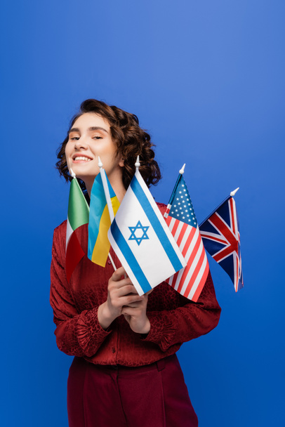 νεαρή εμπνευσμένη γυναίκα που κοιτάζει την κάμερα ενώ κρατά σημαίες από διαφορετικές χώρες που απομονώνονται σε μπλε - Φωτογραφία, εικόνα