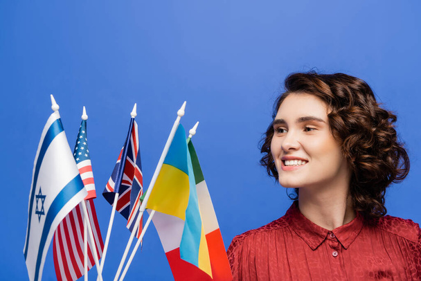 задоволена молода жінка дивиться на прапори різних країн ізольовані на синьому
 - Фото, зображення
