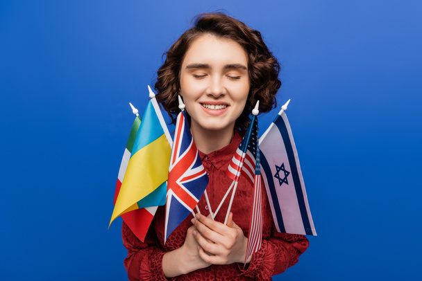 inspired woman with closed eyes holding flags of different countries isolated on blue - Foto, Imagen