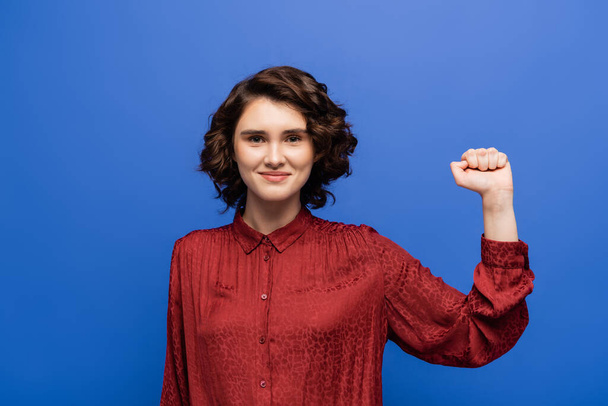 joven mujer alegre en blusa roja gesto de éxito mientras sonríe a la cámara aislada en azul - Foto, imagen