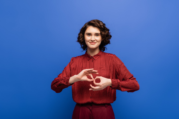 mujer joven y alegre mirando a la cámara y mostrando el símbolo que significa intérprete en el lenguaje de señas aislado en azul - Foto, imagen