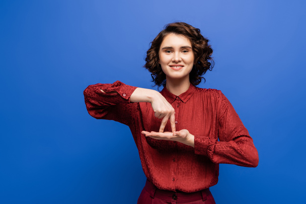 брюнетка женщина улыбается и показывает жест означает стоять на языке жестов изолированы на синий - Фото, изображение