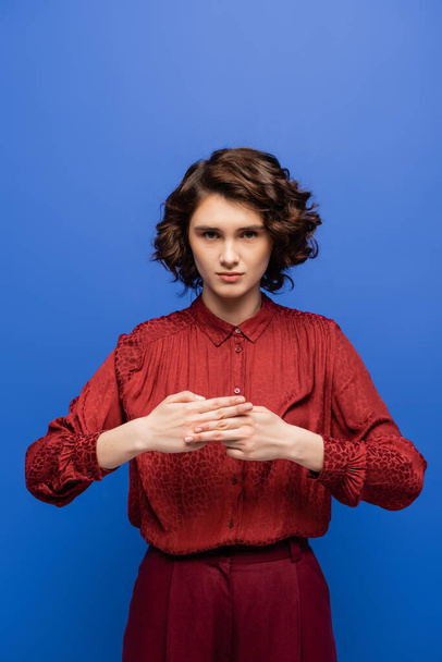 mladá žena s kudrnaté vlasy ukazující znamení znamenající jméno na znakové řeči izolované na modré - Fotografie, Obrázek