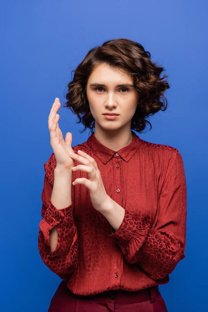 bonita maestra en blusa roja hablando en lenguaje de señas y mirando a la cámara aislada en azul - Foto, Imagen
