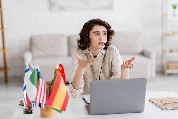 brunette language teacher in headset gesturing near laptop and blurred international flags at home - Foto, Imagem