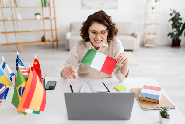 χαρούμενη δασκάλα γλωσσών που δείχνει ιταλική σημαία κατά τη διάρκεια του online μαθήματος σε φορητό υπολογιστή κοντά σε φορητούς υπολογιστές και smartphone  - Φωτογραφία, εικόνα