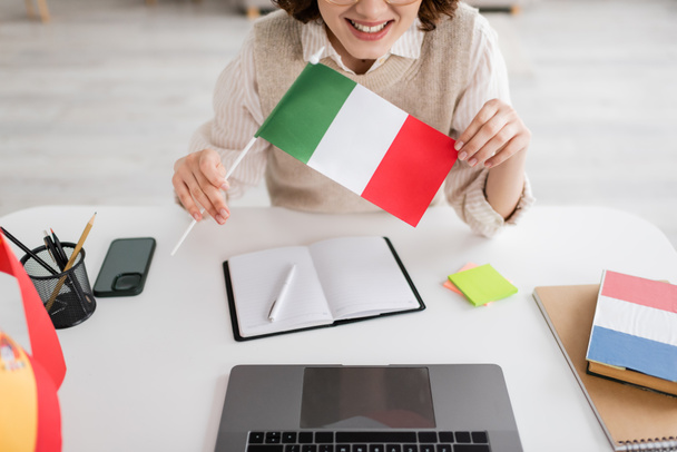 καλλιεργημένη άποψη του χαμογελαστού καθηγητή γλωσσών κρατώντας ιταλική σημαία κοντά σε συσκευές και σημειωματάρια στο τραπέζι στο σπίτι - Φωτογραφία, εικόνα