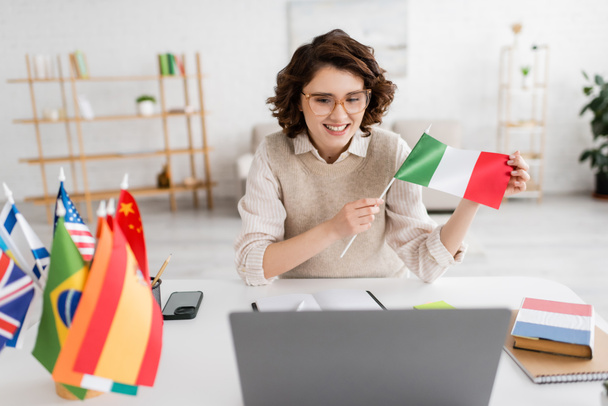 young teacher holding flag of Italy near laptop and textbook of French language while working at home - 写真・画像
