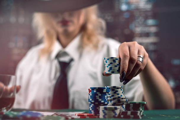 atractiva chica sosteniendo cartas y fichas de póquer en la mesa del casino ocultando su mirada. Jugadora de póquer. casino - Foto, Imagen