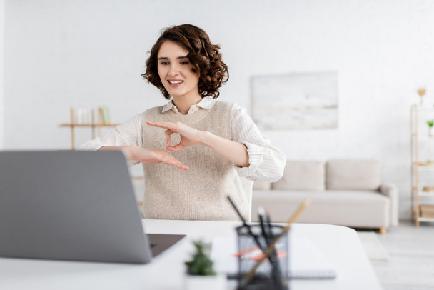 счастливая учительница с кудрявыми волосами, показывающая жест на языке жестов во время онлайн-урока на ноутбуке  - Фото, изображение