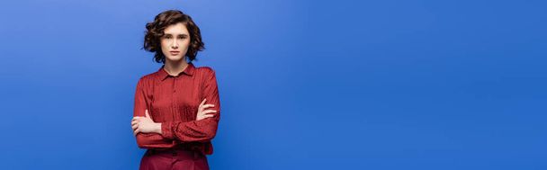 young language teacher in burgundy blouse standing with crossed arms and looking at camera isolated on blue, banner - Foto, Imagem