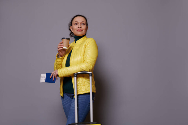 Ελκυστική μεσήλικη πολυεθνική γυναίκα επιβάτης ντυμένη με μπλε τζιν και φωτεινό κίτρινο σακάκι, κοιτάζοντας μακριά στέκεται πάνω από γκρι φόντο με takeaway καφέ σε κύπελλο μιας χρήσης και κάρτα επιβίβασης - Φωτογραφία, εικόνα