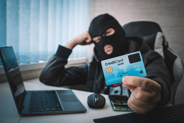 Robo de Internet: un hombre que lleva un pasamontañas y sostiene una tarjeta de crédito mientras está sentado detrás de una computadora portátil, fondo blanco - Foto, imagen