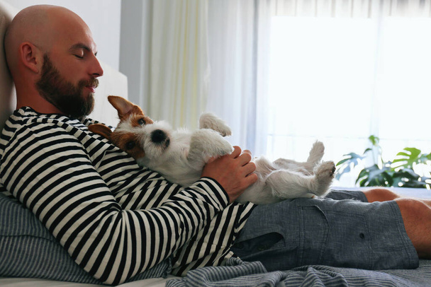 Portrait of a bearded man lying in bed with his wire haired jack russell terrier. Emotional support animal concept. Adult male and his dog napping together. Close up, copy space, background. - Fotografie, Obrázek