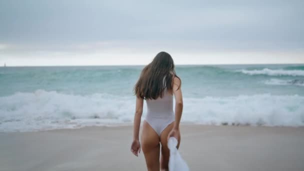 Fırtınalı okyanus dalgaları üzerinde beyaz seksi mayo giymiş uzun saçlı zayıf kadının arka görüntüsü. Bilinmeyen genç kız ıslak kumsal kıyısına basıyor. Baştan çıkarıcı kaygısız bayan bulutlu sahilde yalnız başına rahatla.. - Video, Çekim