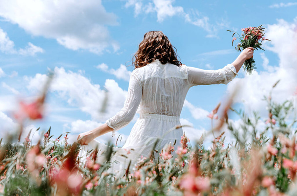 Back view of woman in long white dress with a wildflower bouquet in pink flower field - Zdjęcie, obraz