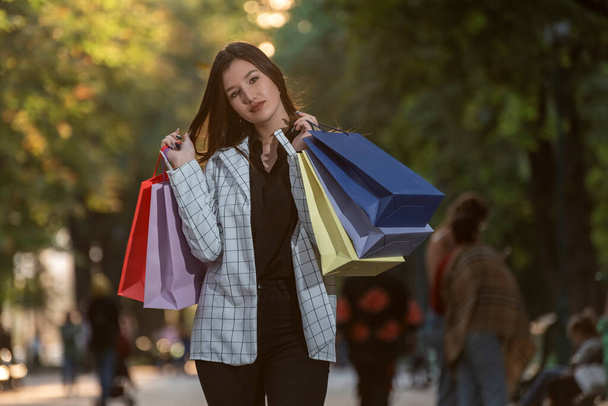 Atrakcyjna stylowa brunetka spaceruje po parku z kolorowymi torbami na zakupy. Dziewczyna z zakupami w rękach - Zdjęcie, obraz