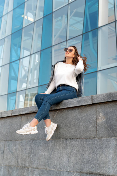 Portrait of stylish young woman in sunglasses on city street. Young girl on background of glass facade of the building - Zdjęcie, obraz