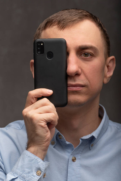 Portrait of man with phone near his face. Man holds black smartphone. Phone instead of concept camera. Vertical frame - Valokuva, kuva