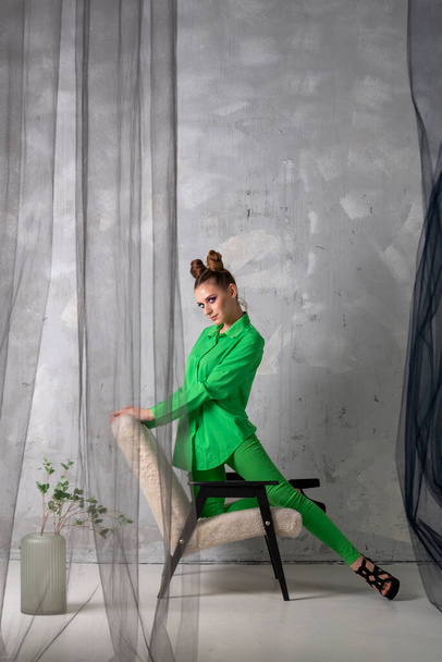 Mooie jonge vrouw met bun kapsel poseert op stoel in de mode studio. Meisje in groen pak en zwarte hakken op grijze muur achtergrond. - Foto, afbeelding