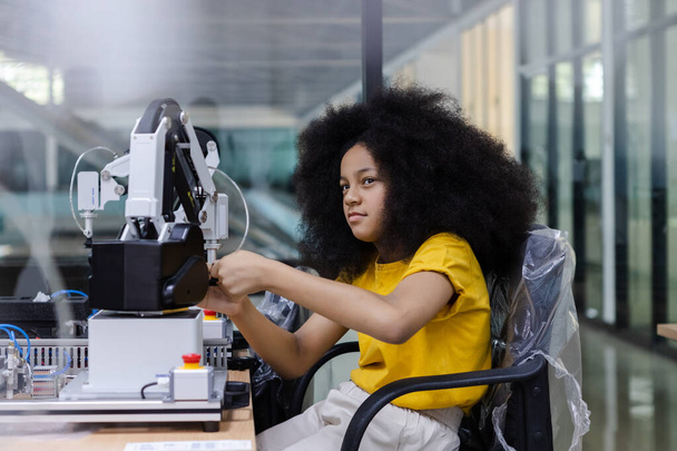 Девушка с африканской причёской носит жёлтую футболку инженера с роботом для обучения на столе в классе. Инновации в робототехнике. - Фото, изображение