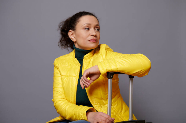 Retrato emocional multiétnico disgustada mujer de mediana edad en una chaqueta de color amarillo brillante, mirando hacia otro lado, expresando emociones tristes, sentado en una maleta sobre un fondo gris aislado con espacio para la publicidad - Foto, Imagen