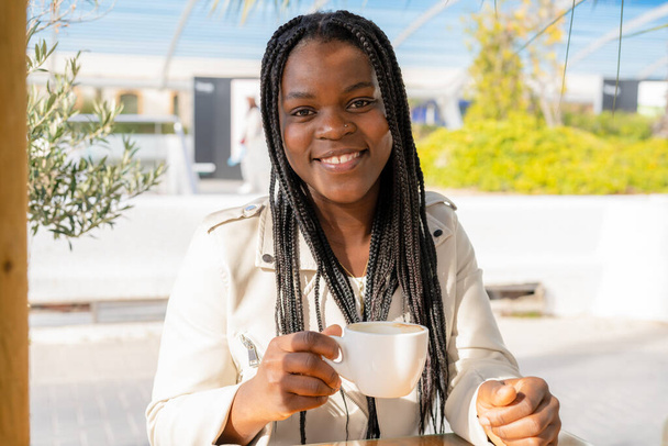 Κοντινό πλάνο μιας γυναίκας με ένα φλιτζάνι καφέ που κάθεται στο καφέ. Αφρικανική γυναίκα που πίνει καφέ σε υπαίθρια καφετέρια. - Φωτογραφία, εικόνα