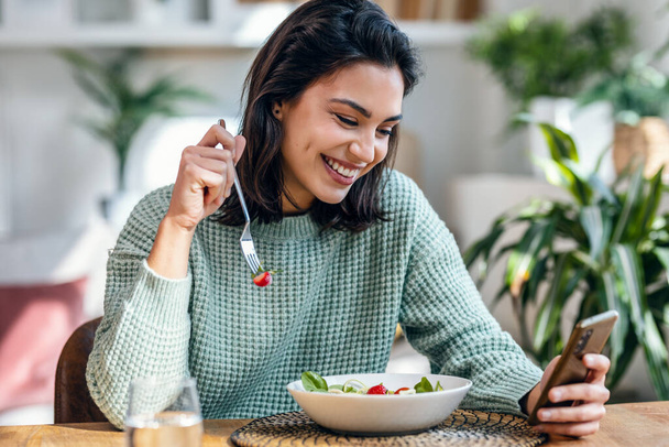 Shot of beautiful young woman eating a bowl of salad while usingh her mobile phone in the living room at home. - Fotografie, Obrázek