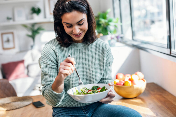Shot of beautiful smiling woman eating healthy salad while sitting on the table at home. - Fotografie, Obrázek