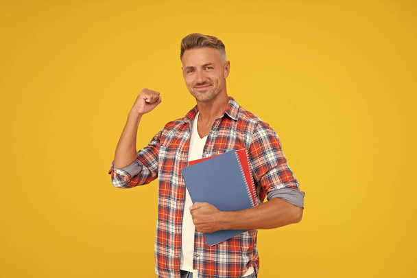 Excited guy teacher making winning gesture holding books yellow background, education. - Photo, Image