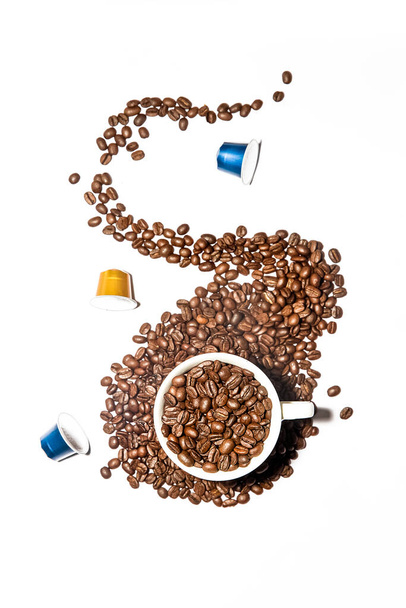 salpicadura de granos de café de una taza blanca llena de granos de café tostados oscuros frescos rodeados con cápsulas de café aisladas sobre fondo blanco. Foto de alta calidad - Foto, Imagen