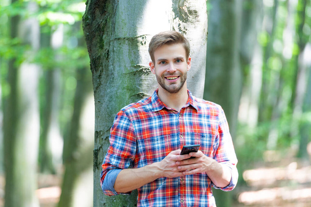 photo of happy man messaging on phone in the forest. man messaging on phone outdoor. man messaging on phone outside. man messaging on phone wearing checkered shirt. - Foto, imagen
