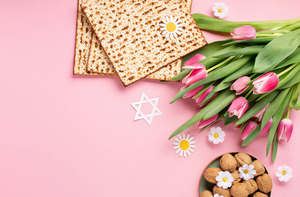 Jewish holiday Passover greeting card concept with matzah, nuts, tulip and daisy flowers on pink table. Seder Pesach spring holiday background, top view, copy space. - Photo, image