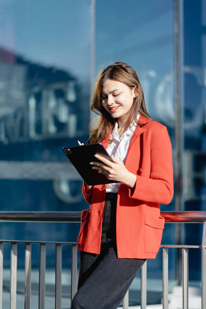 Beautiful woman with long hair holding electronic tablet in front of office buildingYoung successful businesswoman with long brown hair outdoors near building and holding tablet working online - Φωτογραφία, εικόνα