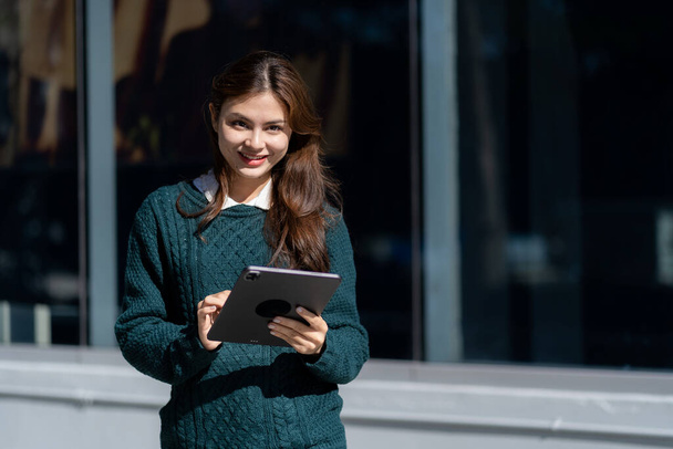 Beautiful woman with long hair holding electronic tablet in front of office buildingYoung successful businesswoman with long brown hair outdoors near building and holding tablet working online - Photo, Image