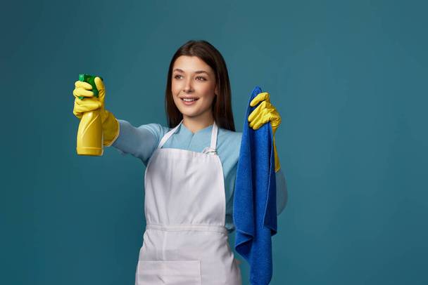 happy young woman in rubber gloves and cleaner apron holding cleaning rag and detergent sprayer on blue background. focus on hands - Φωτογραφία, εικόνα