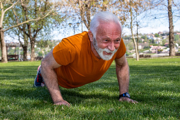 Portrait of a senior man exercising, doing push ups in the park. An elderly man in good shape and health with strong arms. - Foto, afbeelding