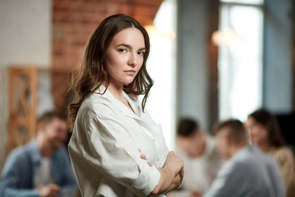 Project manager. Serious ambitious young woman in formal wear attentively looking at camera. Blurred employees on background in office. Concept of business, teamwork, career development, brainstorming - Фото, зображення