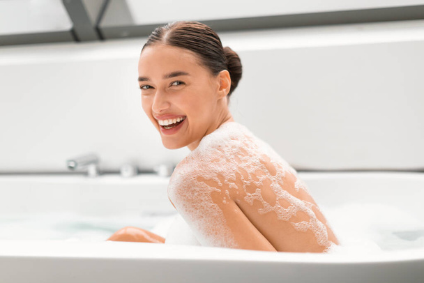 Красивая женщина принимает ванну с пеной улыбаясь глядя на камеру, мытье тела и релаксации сидя в ванной в современной ванной комнате в помещении. Ритуалы красоты и баловство - Фото, изображение