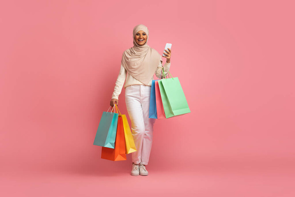 Glimlachende moslimvrouw in hidjab wandelend met smartphone en boodschappentassen in handen, Arabische winkelverslaafde dame met hoofddoek genietend van app met verkoop- en kortingsaanbiedingen, roze achtergrond, kopieerruimte - Foto, afbeelding