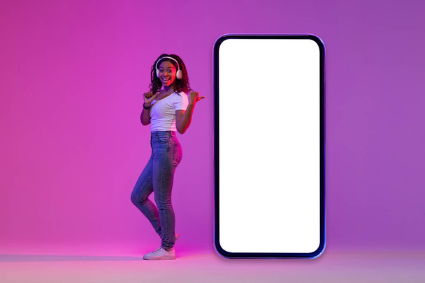 Отличная акция. Веселая черная леди в наушниках, указывающая на большой пустой смартфон с белым экраном, счастливая афроамериканская женщина, стоящая в неоновом свете на фиолетовом фоне, коллаж, макет - Фото, изображение