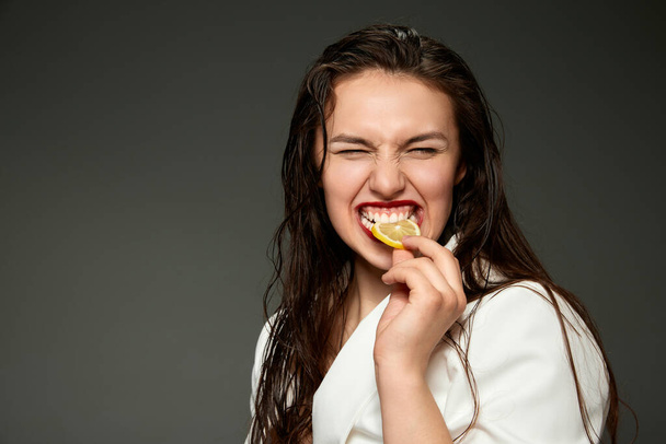 Funny whitening. Emotional fashionable stylish beautiful woman wearing white jacket tasting slice of orange over gray background. Concept of healthy lifestyle, vitamins, dieting, fashion, beauty - Fotoğraf, Görsel