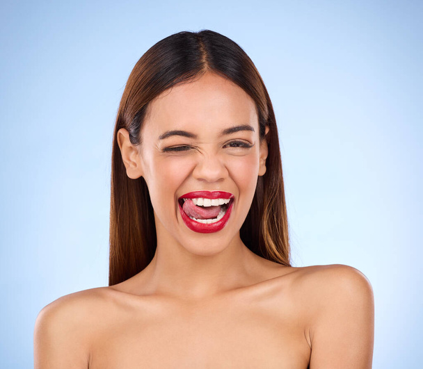 Wink, woman and red lipstick makeup portrait with cosmetics on face in studio. Aesthetic female model on a blue background for self care, facial glow and beauty or color for skin with tongue out. - Photo, image