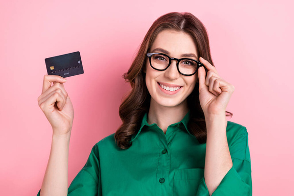 Photo of toothy beaming woman with wavy hairdo dressed green shirt touching eyewear holding plastic card isolated on pink background. - Fotoğraf, Görsel