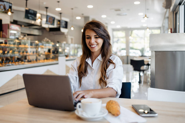 Beautiful and happy young woman sitting and eating delicious rolls in bakery or fast food while she using laptop computer. - Фото, зображення