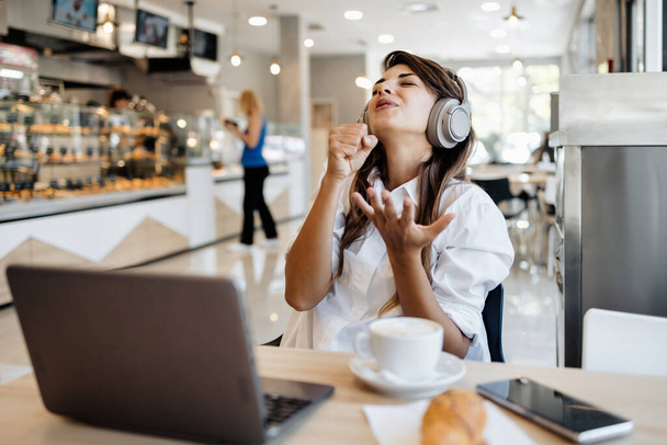 Beautiful and happy young woman sitting and eating delicious rolls in bakery or fast food. She also using her laptop and listening to music with headphones.  - Foto, Bild
