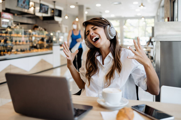Beautiful and happy young woman sitting and eating delicious rolls in bakery or fast food. She also using her laptop and listening to music with headphones.  - Foto, Bild